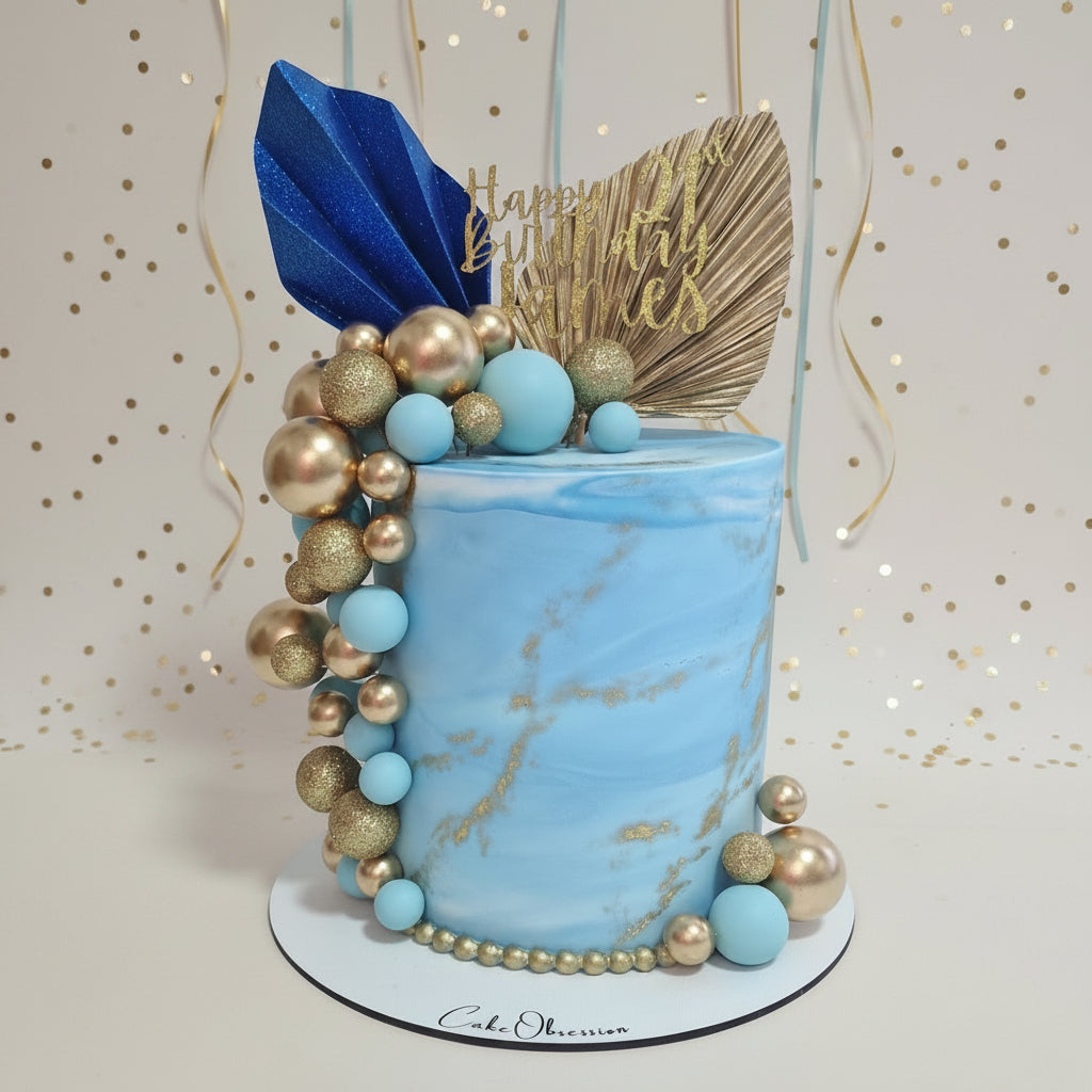 A decorated cylindrical birthday cake with a light blue marble finish, topped with gold and blue decorative elements including a blue leaf topper, gold fan topper, and gold 'Happy Birthday' script. The cake is decorated with a cascading arrangement of light blue and metallic gold beads on a white round board.