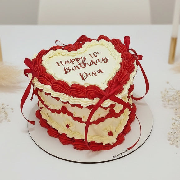 A heart-shaped birthday cake with red and white piped frosting, decorated with red ribbons and a white center card that reads 'Happy Birthday Ben', shown on a white round board.