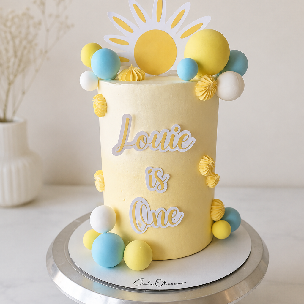 Sunny yellow, blue and white birthday cake with ombre buttercream, cake balls and personalised name, available from CakeObsession in Essex.