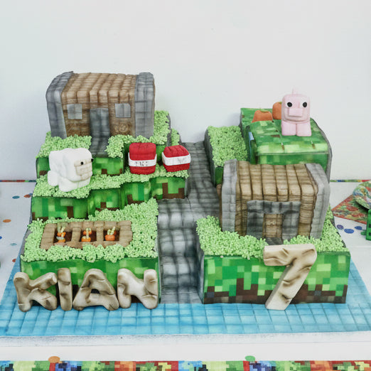 A themed cake designed to look like a Minecraft village, featuring green grass, brown buildings, and a pink pig. Minecraft village themed birthday cake made in Basildon Essex for kids celebration