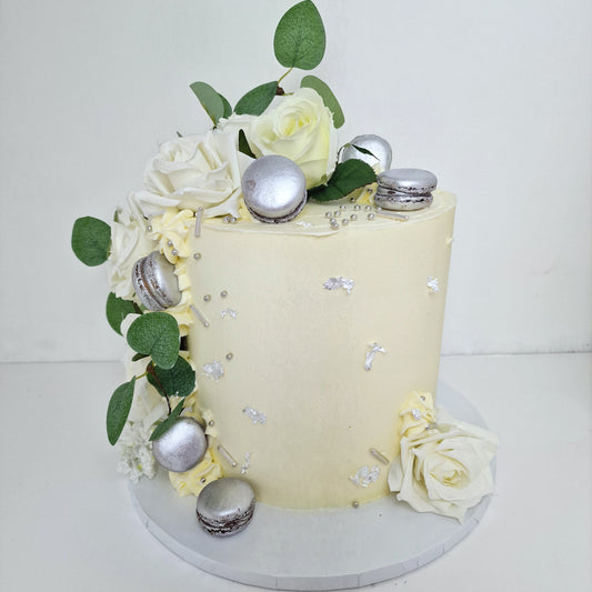 Indulge in our simple yet elegant cake, adorned with silk white roses, edible silver or gold accents, and topped with delightful macarons. This exquisite creation is perfect for any special occasion.