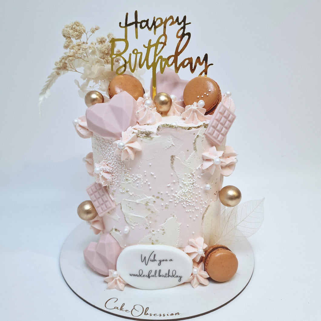 Indulge in a beautiful birthday cake adorned with soft pink buttercream, topped with elegant dried flowers and delectable macaroons. The color of the macaroons and dried flowers may vary based on stock availability, ensuring a unique and personalized touch to each cake. Perfect for making any celebration extra special.
