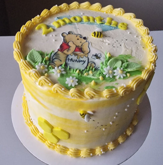 A round birthday cake decorated with yellow frosting, piped borders, green leaves, white flowers, and a cartoon bear character on top, held on a white board by a hand against a plain gray background.