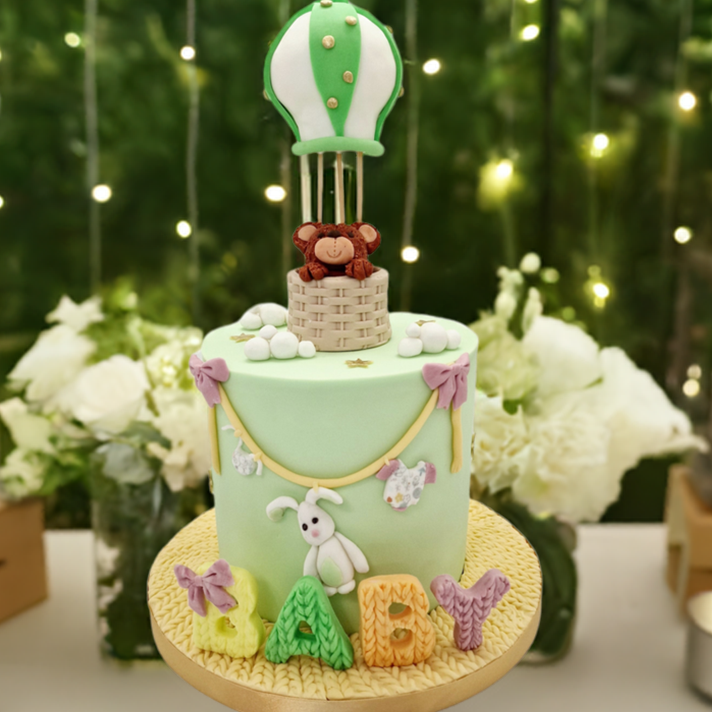 A light green baby shower cake with a teddy bear inside a hot air balloon, decorated with textured letters spelling 'baby', placed on a table with candles and decorative elements in the background.