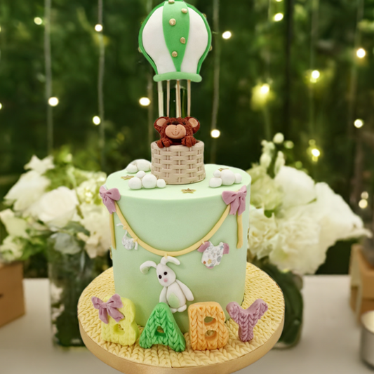 A light green baby shower cake with a teddy bear inside a hot air balloon, decorated with textured letters spelling 'baby', placed on a table with candles and decorative elements in the background.