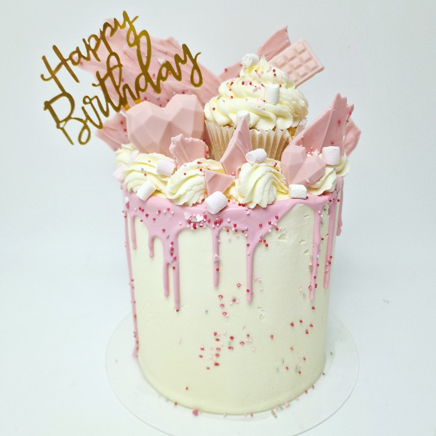 A decorated birthday cake with white frosting and pink chocolate drips, topped with pink cupcake shards and a 'Happy Birthday' text topper.