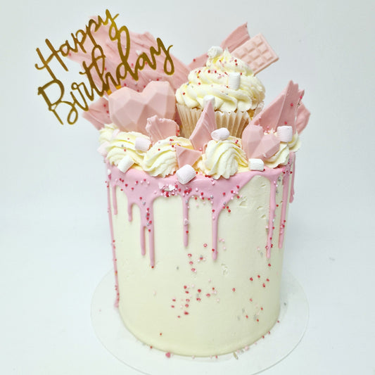 A decorated birthday cake with white frosting and pink chocolate drips, topped with pink cupcake shards and a 'Happy Birthday' text topper.