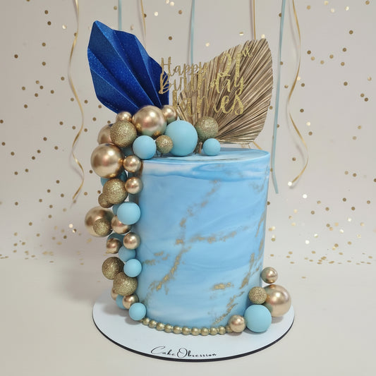 A decorated cylindrical birthday cake with a light blue marble finish, topped with gold and blue decorative elements including a blue leaf topper, gold fan topper, and gold 'Happy Birthday' script. The cake is decorated with a cascading arrangement of light blue and metallic gold beads on a white round board.