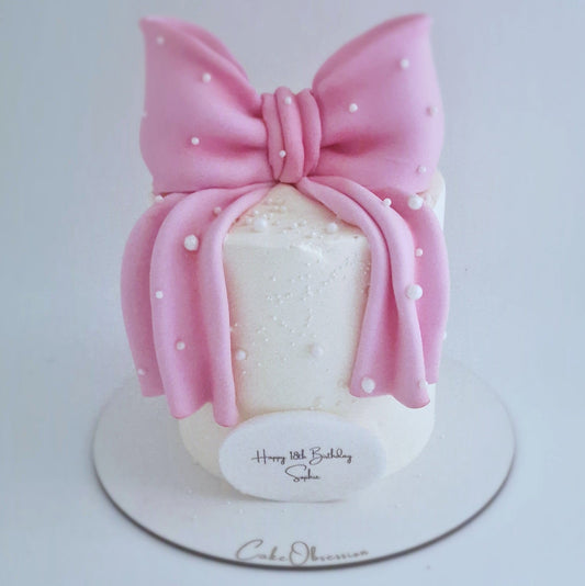 White buttercream cake decorated with a large pink fondant bow and pearl details, displayed on a cake board.