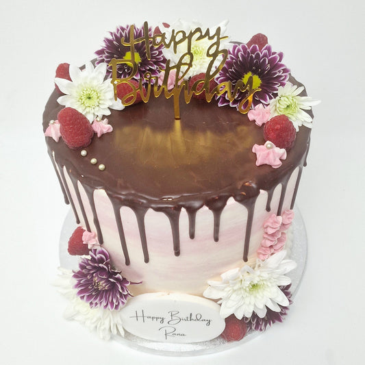 A beautifully decorated white buttercream cake with a pink chocolate drip, topped with fresh berries and delicate flowers, perfect for elegant celebrations.