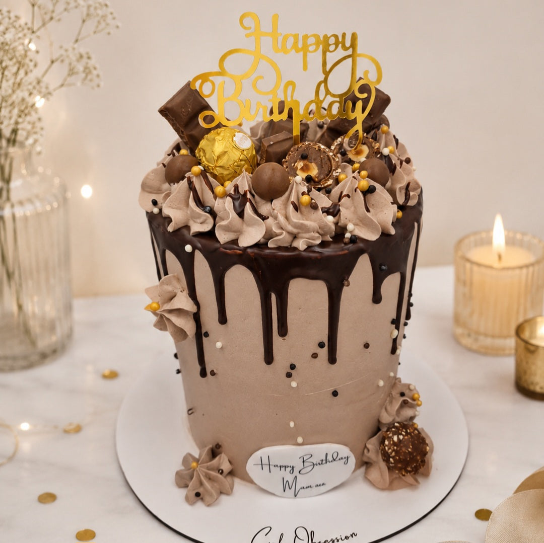 A decorated birthday cake with chocolate drip frosting, piped cream frosting, chocolate-covered strawberries, and gold and brown chocolates, topped with a gold 'Happy Birthday' sign and a small 'Happy Birthday Mama' plaque on a white board.