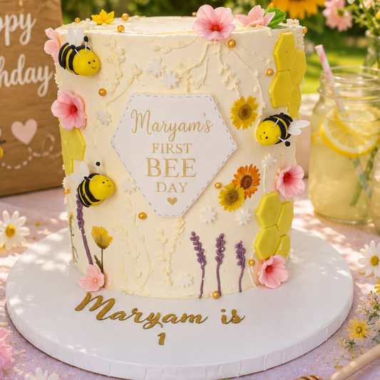 A round white frosted birthday cake decorated with yellow honeycomb shapes, pink flowers, bumblebee ornaments, and purple lavender stems, with a central hexagon plaque reading 'Maryam's FIRST BEE DAY.' The cake sits on a white round board with gold script text reading 'Maryam is 1,' photographed in a bright outdoor party setting with flowers and a drink jar in the background.