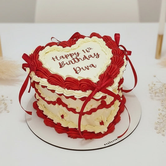 A heart-shaped birthday cake with red and white piped frosting, decorated with red ribbons and a white center card that reads 'Happy Birthday Ben', shown on a white round board.