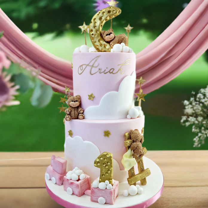 A decorated first birthday cake with pink fondant, golden moon and stars, teddy bears, a toy train, and white clouds, set on a wooden table outdoors with a floral backdrop. First birthday cake made in Basildon Essex for baby girl celebration