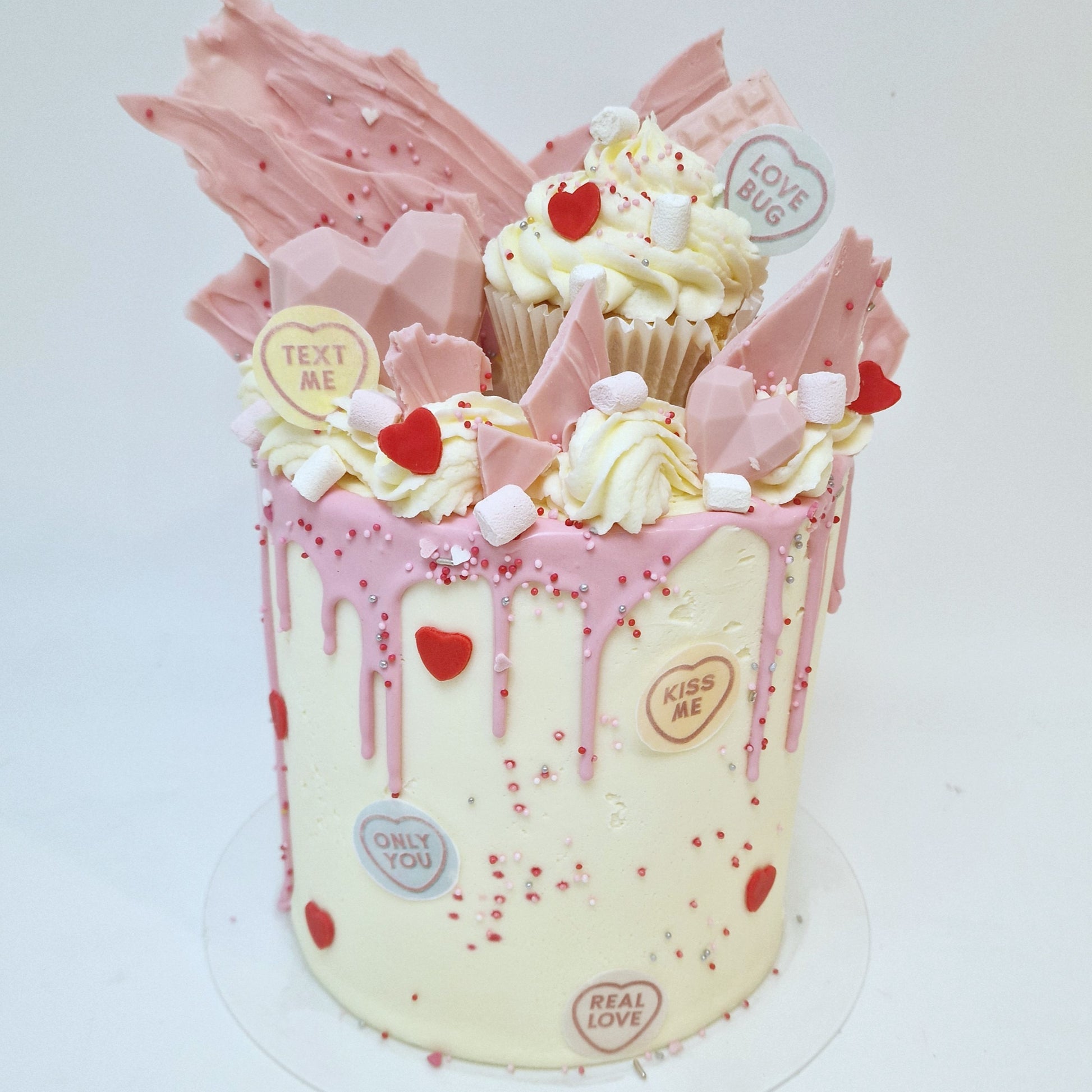 A beautifully decorated Valentine’s Day cake with a smooth pink drip cascading down the sides. The cake is topped with heart-shaped decorations, delicate swirls of frosting, and sprinkles, evoking a romantic and festive theme. The base color of the cake is light, complementing the vibrant pink drip.