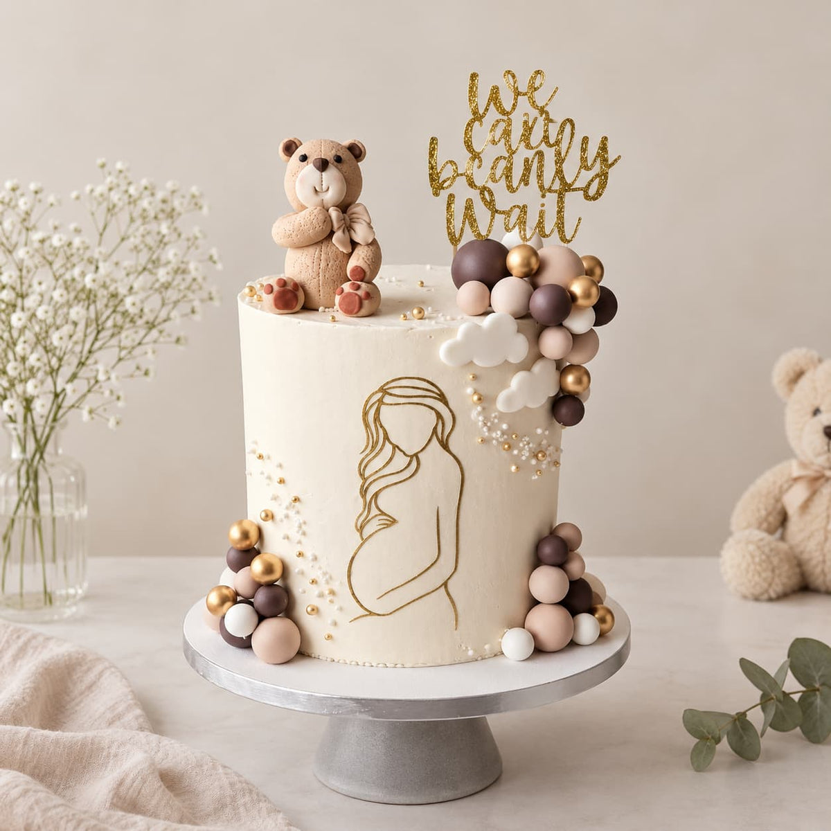 A decorated round cake on a silver pedestal stand, featuring a gold line drawing of a pregnant figure, pastel balloon clusters, a small teddy bear topper, and a gold script topper reading 'we can be ready wait'. The scene is styled with baby flowers, a folded cream cloth, and a teddy bear on a light neutral background.