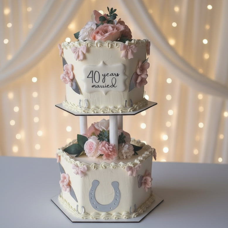 A two-tiered cream-colored wedding anniversary cake on a white pedestal stand. The top tier is decorated with pink roses, green leaves, and a white plaque that reads '40 years married'.