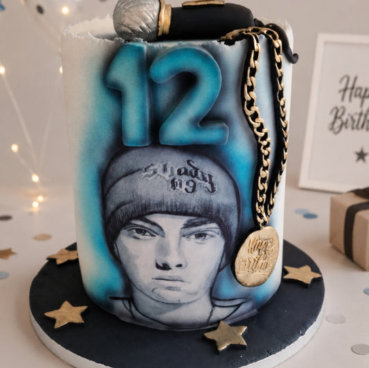A custom birthday cake decorated with a portrait of Eminem wearing a beanie, blue and white gradient shading, a gold chain with a medallion, and a microphone on top, set on a black base with gold star accents.