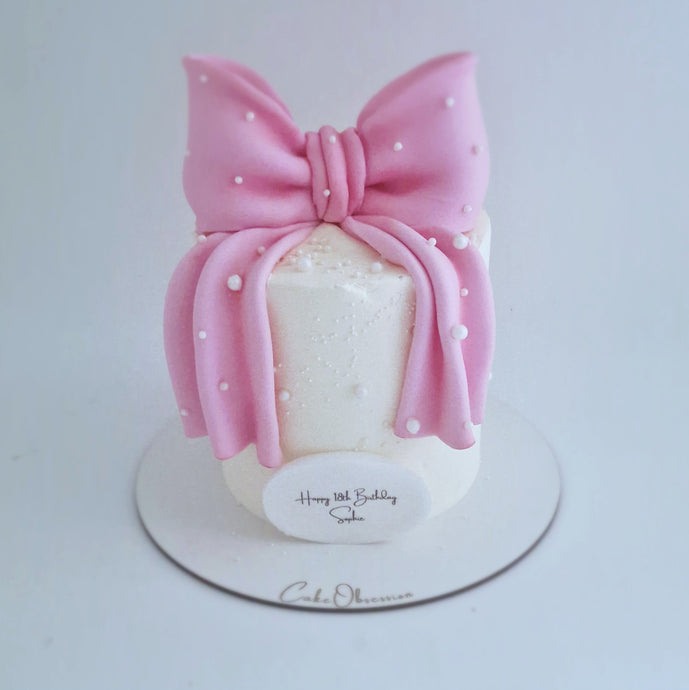 White buttercream cake decorated with a large pink fondant bow and pearl details, displayed on a cake board.