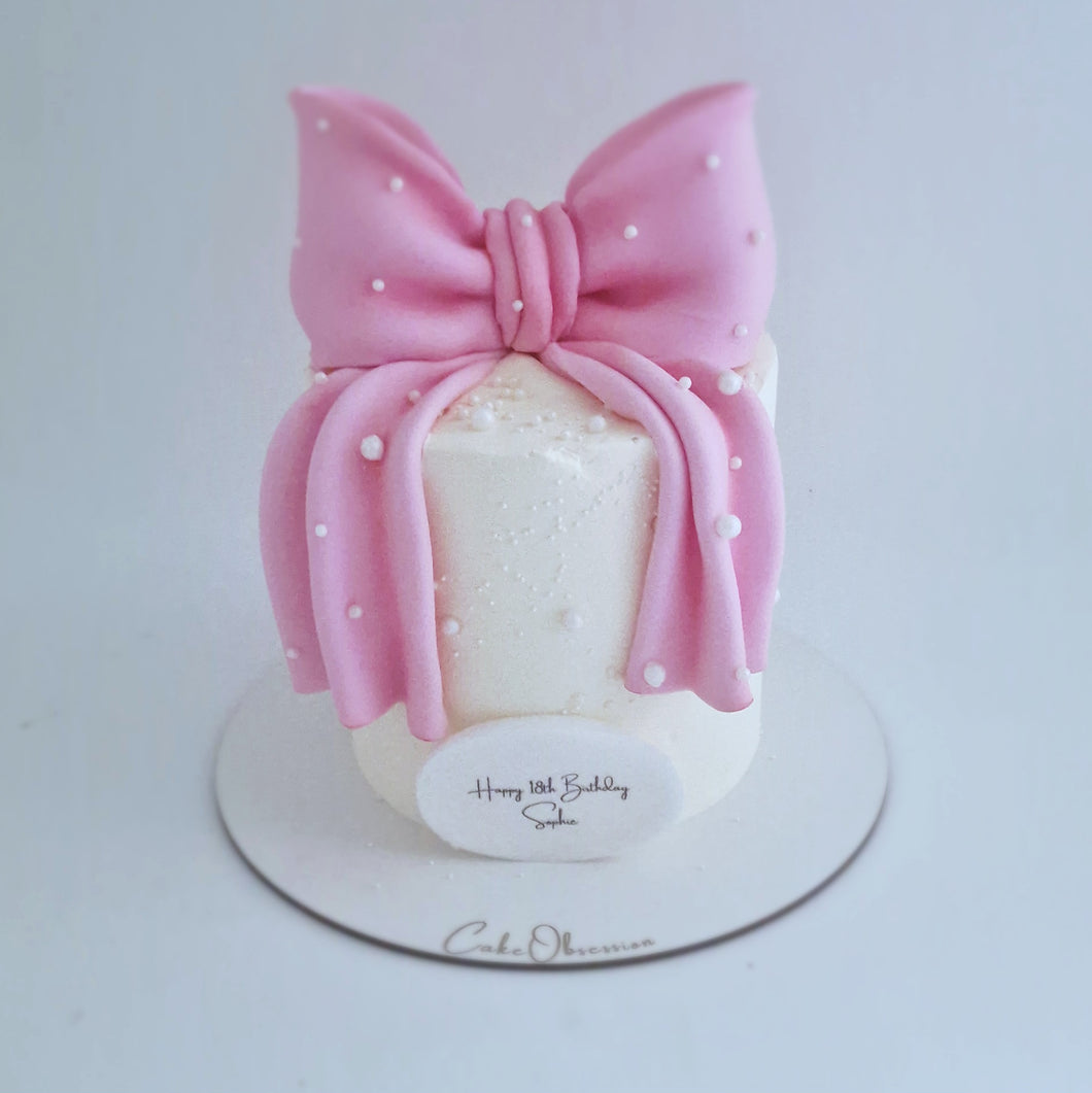 White buttercream cake decorated with a large pink fondant bow and pearl details, displayed on a cake board.