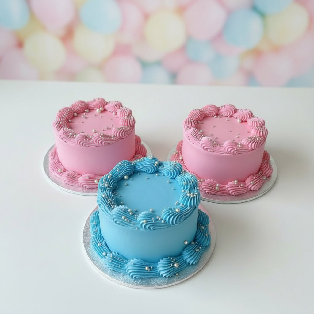 Three small personalized birthday cakes with pink and blue frosting, gold script names, and gold sprinkles, displayed on a gray woven table outdoors.