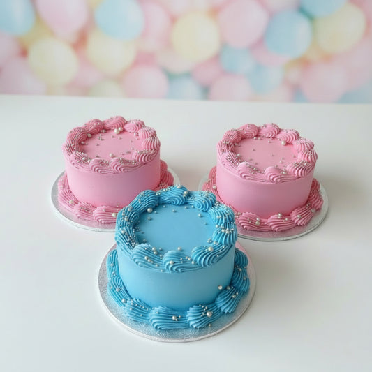 Three small personalized birthday cakes with pink and blue frosting, gold script names, and gold sprinkles, displayed on a gray woven table outdoors.