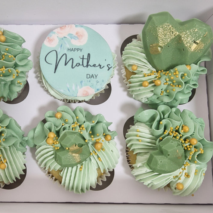 Green cupcakes with gold decorations in a box, one with a 'Happy Mother's Day' card.