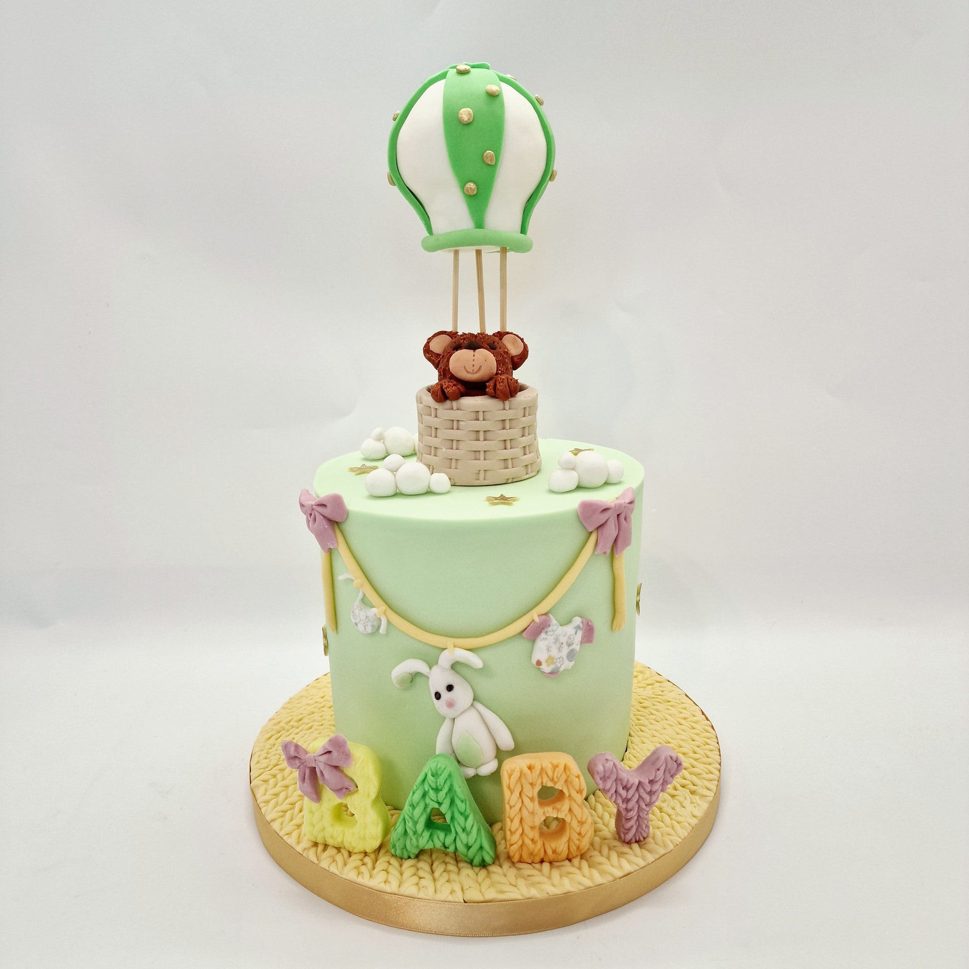 A light green baby shower cake with a teddy bear inside a hot air balloon, decorated with textured letters spelling 'baby', placed on a table with candles and decorative elements in the background.